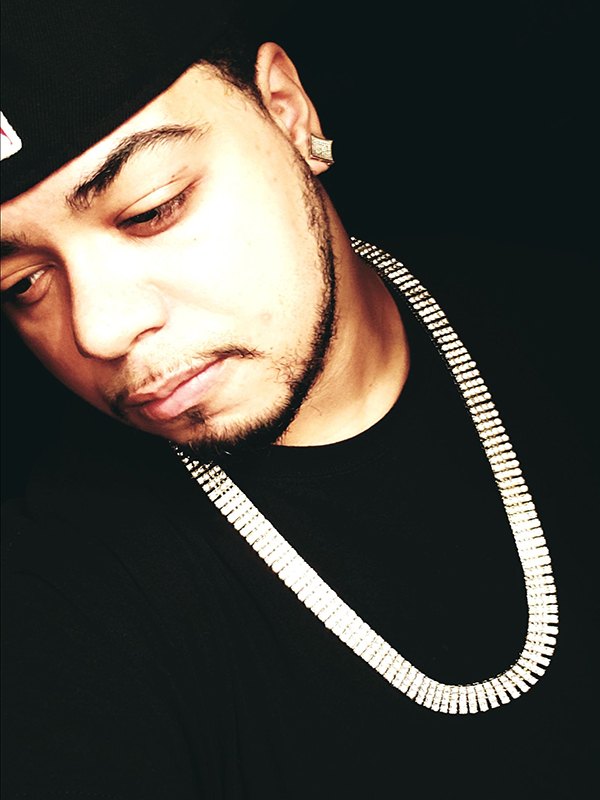 XiØN in a black cap and black shirt, wearing a silver cuban link chain against a dark studio background